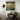 A framed abstract painting with earthy green, beige, and brown tones hangs on a light wall above a brown armchair with cushions and a draped green throw. A tall amber floor lamp stands to the left.