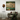 An abstract framed painting with green, beige, and brown tones hangs on a light gray wall above a brown leather armchair with neutral cushions and a draped green throw. A brown floor lamp stands to the left.