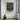 A framed abstract landscape painting hangs on a white wall in a modern room setting, featuring a textured green and beige design. The frame is dark brown, and the painting is styled in a minimalist interior with a dark ceramic vase on the wooden floor below.