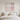 A minimalist bedroom scene with a large square abstract wall art piece hanging above a neatly made bed. The artwork features soft vertical gradients in blush pink, cream, and light gray, framed in a thin light wood border.