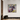 A square framed abstract painting with expressive brushstrokes in purple, yellow, green, white, and pink, hanging on a white wall in a modern interior setting with a dark wooden cabinet and a black textured planter.