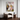A framed abstract floral painting with pink, white, green, and purple brushstrokes hangs on a white wall above a modern wooden armchair and a small round side table with a globe.