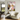 A large framed abstract canvas print with pink, white, purple, and yellow brushstrokes hangs on a light gray wall in a modern living room setting next to a cream-colored armchair and a small black side table.